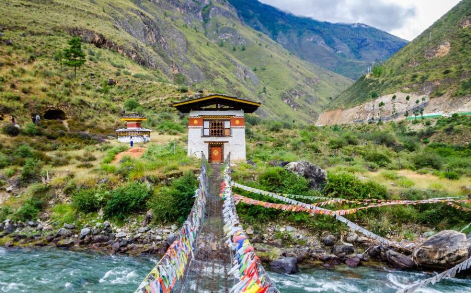 Customs and traditions in Bhutan - Iron Chain Bridge of Tamchog Lhakhang Monaster over the Paro River in Bhutan. Colourful flags adorn the sides of the wooden swinging bridge and towards a small hut-like building, and mountains sit in the backdrop of the photo.